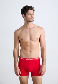 Homme torse nu avec des cheveux foncés portant des boxers rouges Björn Borg, se tenant contre un fond clair uni, regardant sur le côté.