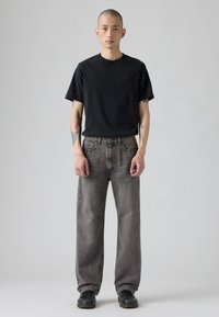 Crna majica s okruglim vratom u kombinaciji sa svijetlosivim hlačama ravnog kroja. Model nosi crnu obuću. Jednostavan dizajn, bez vidljivih uzoraka ili grafika.