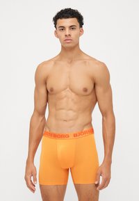 Boxer shorts laranja feitos de um tecido elástico com uma cintura ajustável apresentando a marca "BJÖRN BORG" em vermelho. Textura suave e design ajustado.