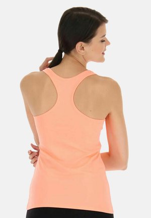 Vrouw met donker haar in een paardenstaart, gekleed in een mouwloos perzikkleurig racerback-topje, staand met handen achter de rug, zijwaarts gericht.