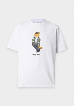 Ung mand iført hvid t-shirt med tegneserie-ræv, blå jeans, sort bælte og hvide sneakers stående med hænderne i lommerne.