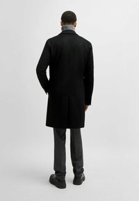 Cappotto di lana nero con colletto alto e colletto interno grigio testurizzato; abbinato a pantaloni grigi e scarpe nere. Design semplice e strutturato.