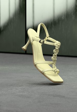 Blekgul høyhælte sandal med stropper og blomsterdetaljer som svever over en grå gulvflate og reflekterende mørk veggbakgrunn.