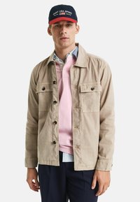 Beige leichte Jacke mit zwei Brusttaschen und Knöpfen; über einem pinken Poloshirt und einem blau karierten Hemd getragen; kombiniert mit dunklen Hosen.