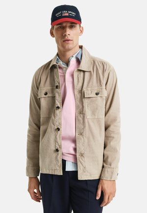 Veste légère beige avec deux poches poitrine et des boutons ; superposée sur un polo rose et une chemise à carreaux bleue ; associée à un pantalon sombre.
