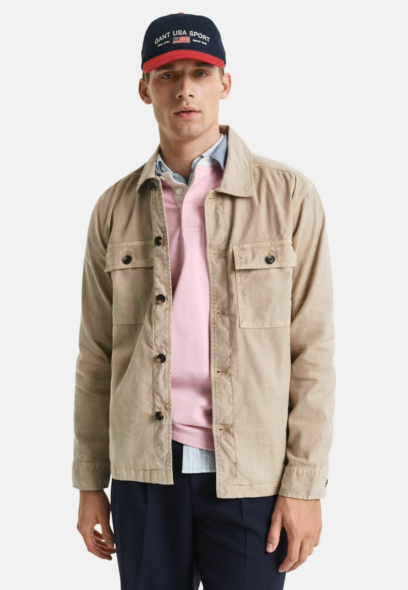 Beige leichte Jacke mit zwei Brusttaschen und Knöpfen; über einem pinken Poloshirt und einem blau karierten Hemd getragen; kombiniert mit dunklen Hosen.
