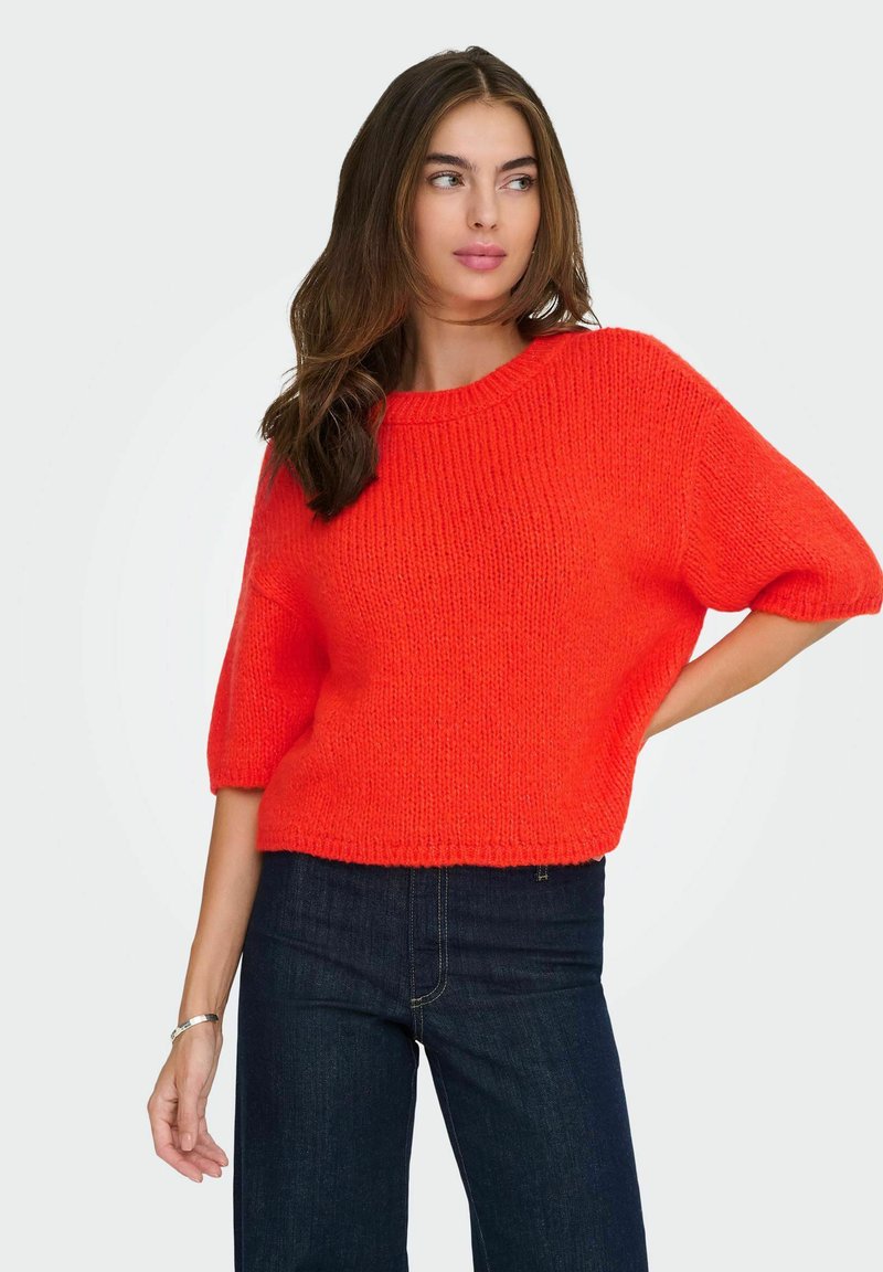 Pull en tricot de couleur orange vif, avec un col rond, des manches courtes et une coupe ample. Assorti à un jean en denim foncé.