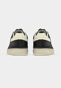 Paire de baskets basses noires et blanches vues de dos avec des semelles beiges texturées sur un fond clair uni.