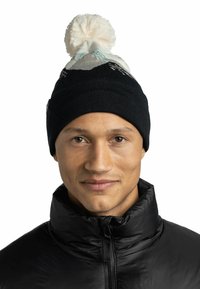 Gorro de punto negro con un pompón blanco, que presenta un patrón de montañas en azul claro y negro. Se lleva con una chaqueta de plumas negra.