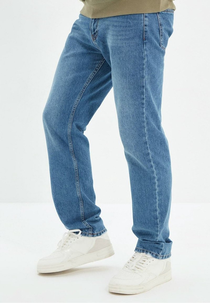 Jeans in denim blu chiaro con taglio dritto e lievi dettagli in whiskering. Indossati con sneakers bianche con lacci su uno sfondo semplice.