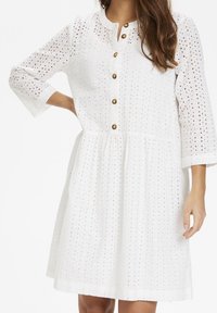 Weißes Kleid mit Lochmuster, Dreiviertelärmeln, runder Ausschnitt und bronzefarbenem Knopfverschluss vorne. A-Linie Silhouette.