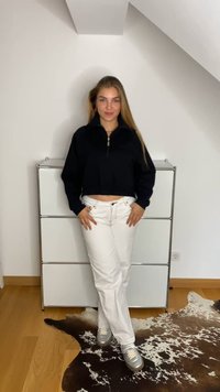 Pullover preto cropped com gola alta, combinado com jeans brancos. Ténis cinzentos completam o look. O ambiente apresenta um chão de madeira clara.