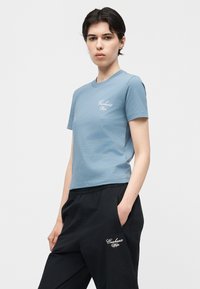 Νέος που φοράει μπλε μπλουζάκι Carhartt Wip και μαύρο παντελόνι Carhartt Wip, στέκεται με το ένα χέρι στην τσέπη μπροστά σε απλό φόντο.