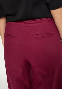Bordowe spodnie o gładkiej fakturze, z dwoma tylnymi kieszeniami i dopasowanym krojem w talii. Brak widocznych zapięć.