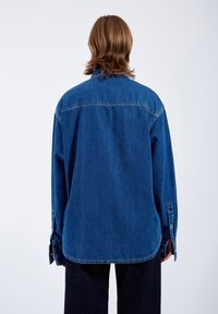 Camicia in denim di colore blu scuro, taglio rilassato, con maniche lunghe, polsini a bottone e colletto a punta. Cuciture visibili e texture liscia.