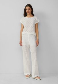 Weißes Baumwolloberteil mit Durchbruchsdesign und kurzen Ärmeln, kombiniert mit weißen, locker sitzenden Hosen mit gesticktem Blumendesign. Weiße Sandalen.
