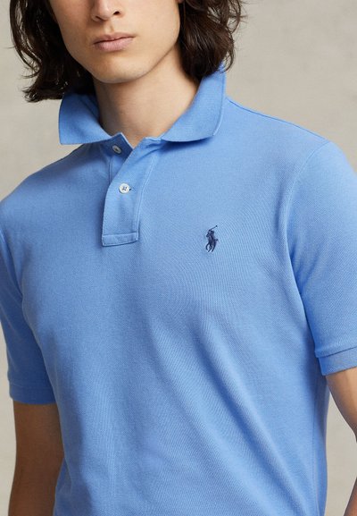Jaunas asmuo dėvi šviesiai mėlyną polo marškinėlį su mažu tamsiu siuvinėtu arkliu ir raiteliu ant krūtinės.