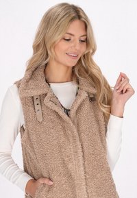 Beige Fleece-Weste mit strukturiertem Oberflächen, Kragen und Druckknöpfen. Darunter ein weißes Langarmshirt sowie eine sichtbare geschichtete Halskette.