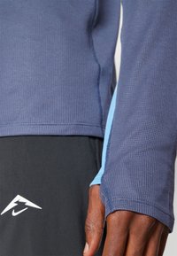 Haut à manches longues texturé bleu marine avec des accents bleu clair, présentant un design sans couture le long des manches et un petit logo blanc sur la poitrine.