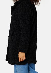 Schwarzer, flauschiger, strukturierter Mantel mit lockerer Passform, seitlichen Taschen und Reverskragen. Über blauen Jeans getragen, im Profil dargestellt.