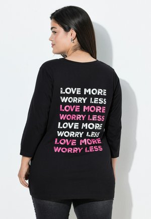 Svart långärmad t-shirt med den upprepade texten "ÄLSKA MER Oroa DIG MINDRE" i olika typsnitt och färger, inklusive rosa och vitt, på baksidan.
