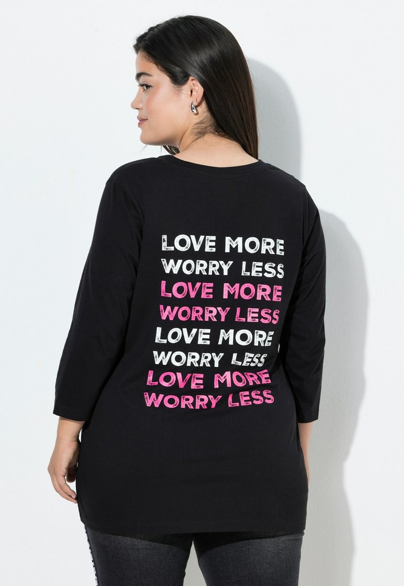 Svart långärmad t-shirt med den upprepade texten "ÄLSKA MER Oroa DIG MINDRE" i olika typsnitt och färger, inklusive rosa och vitt, på baksidan.