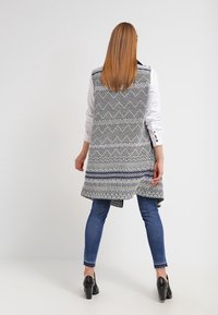 Gilet lungo e lavorato in bianco e nero con design geometrici, caratterizzato da un orlo asimmetrico, indossato sopra una camicia bianca e jeans blu.