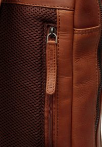 Bolso de cuero marrón con un panel de malla, cierre de cremallera y un acabado texturizado. Incluye una etiqueta de cuero decorativa con texto embossado.