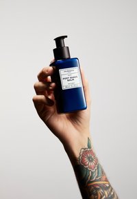 Niebieska szklana butelka z czarną pompką, opisana białym napisem "Balsam po goleniu", trzymana w ręce z kolorowymi tatuażami na nadgarstku.