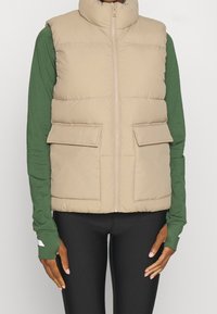 Veste matelassée beige avec col montant, fermeture éclair frontale et deux poches à rabat. Portée sur un chemisier vert à manches longues avec des leggings noirs ajustés.