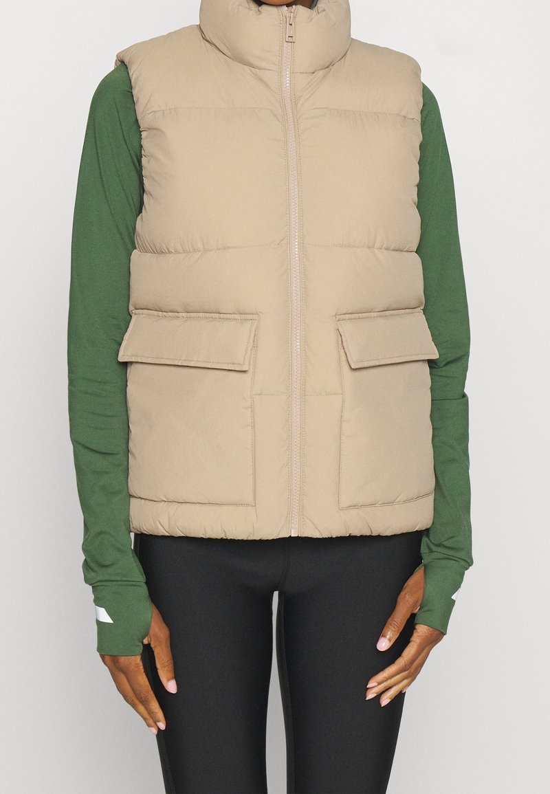 Veste matelassée beige avec col montant, fermeture éclair frontale et deux poches à rabat. Portée sur un chemisier vert à manches longues avec des leggings noirs ajustés.