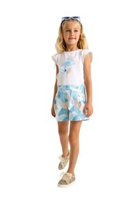 Camicetta bianca a maniche corte con design floreale blu, abbinata a pantaloni corti azzurri con fantasia. La ragazza indossa sneakers dorate glitterate con fiocchi.