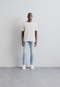 Ανοιχτό μπεζ μπλουζάκι, εφαρμοστά ανοιχτό γαλάζια τζιν, λευκά αθλητικά παπουτσάκια. Το μοντέλο στέκεται μπροστά από γκρίζο φόντο, με τα χέρια χαλαρά στα πλάγια.