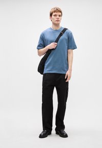 Μπλε βαμβακερό t-shirt με κοντά μανίκια, συνδυασμένο με μαύρο cargo παντελόνι που έχει τσέπες με φερμουάρ και μαύρα υποδήματα. Το μοντέλο κρατά μια μαύρη τσάντα ώμου.