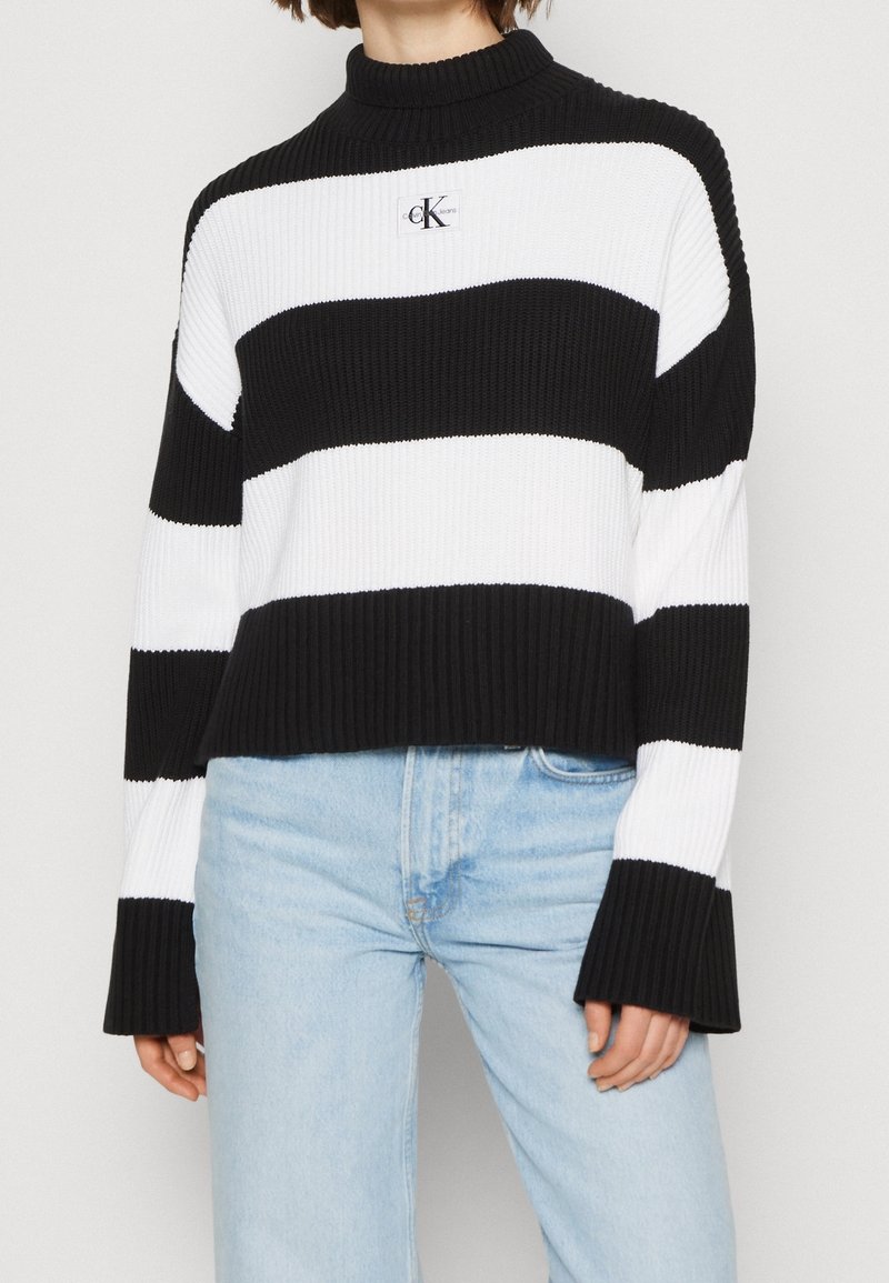 Sweter dzianinowy w czarno-białe paski z wysokim kołnierzem, prążkowaną fakturą i naszywką z logo na piersiach. Rękawy są szerokie i dzwoniaste.