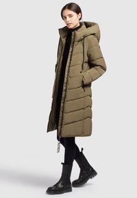 Young woman wearing an olive green quilted long coat with hood, black leggings, and black ankle boots, standing against a plain light background.