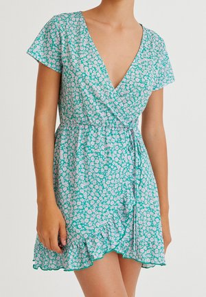 Frau trägt ein kurzes grünes Wickelkleid mit weißem Blumenmuster, kurzen Ärmeln, Rüschensaum und gebundenem Taillengürtel.