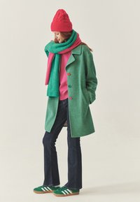 Grøn sildebensmønstret frakke med farverige knapper, lyserød sweater og tørklæde. Mørkeblå vide jeans og grønne sneakers med hvide striber fuldender outfittet.