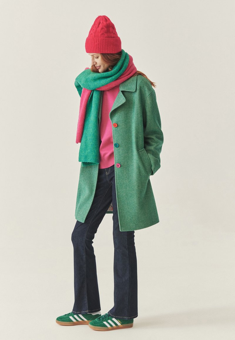 Grøn sildebensmønstret frakke med farverige knapper, lyserød sweater og tørklæde. Mørkeblå vide jeans og grønne sneakers med hvide striber fuldender outfittet.