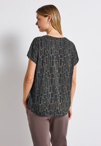 Top a maniche corte di colore verde scuro con un motivo astratto beige, caratterizzato da un fit rilassato e un tessuto morbido e leggero. Dettagli di cucitura visibili.