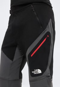 Nahaufnahme von schwarzen und grauen Outdoor-Hosen mit roter Reißverschlusstasche und The North Face-Logo am Oberschenkel.