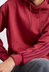 Vinröd hoodie med känguruficka, ribbade ärmslut och huva med dragsko. Har tre lila ränder på varje ärm och en präglad logotyp.