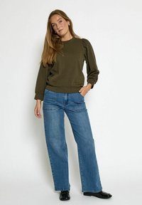Oliwkowy sweter z bufiastymi rękawami, zestawiony z jasnoniebieskimi spodniami w szerokim kroju. Model stoi na neutralnym tle. Czerny obuwie.