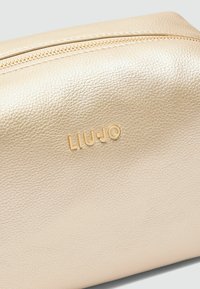 Zlata umetna kožna toaletna torbica z teksturirano površino, zadrgo in vtisnjenim logotipom "LIU·JO" na sprednji strani. Okrogla oblika.
