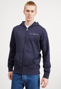 Felpa zip-up blu navy in misto cotone, dotata di tasca a marsupio, polsini a costine e logo GANT 1949 sul petto. Vestibilità casual.