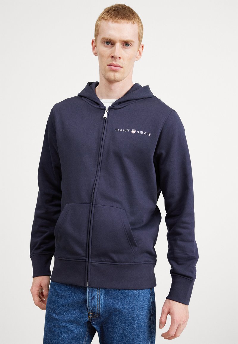 Felpa zip-up blu navy in misto cotone, dotata di tasca a marsupio, polsini a costine e logo GANT 1949 sul petto. Vestibilità casual.