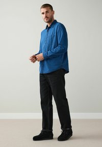 Blå skjorta med knappar och texturerad yta, bröstficka, kombinerad med svarta jeans och svarta snörskor mot en neutral bakgrund.