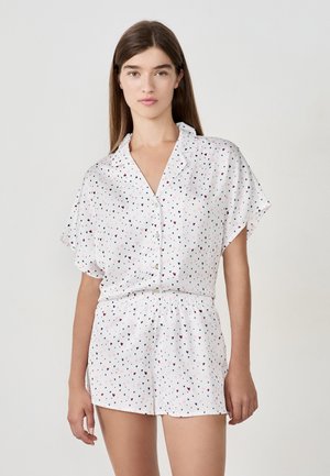 Jeune femme portant un haut blanc à manches courtes avec des boutons et un short assorti, orné de petits motifs en forme de cœurs noirs, roses et bordeaux.