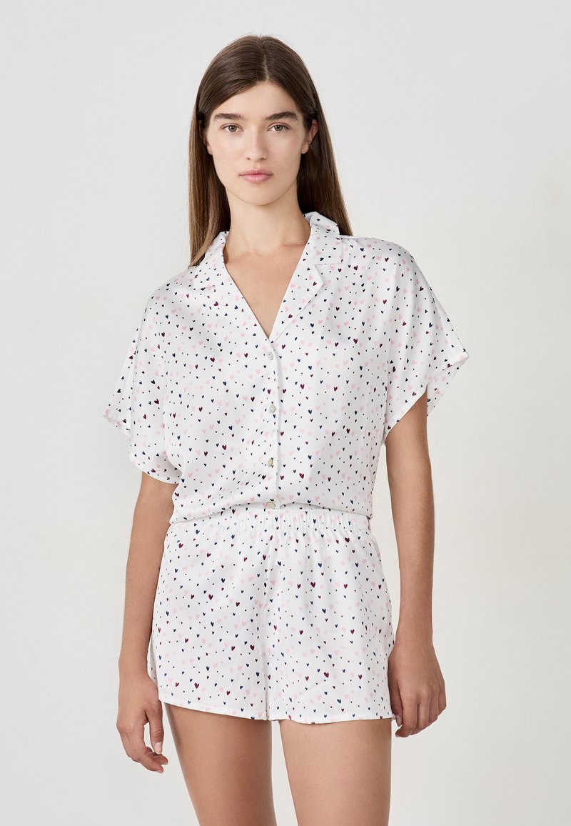 Joven mujer vistiendo una camisa blanca de botón y manga corta, junto con pantalones cortos a juego con pequeños patrones de corazones en negro, rosa y burdeos.