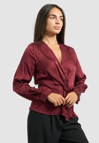 Blusa di seta bordeaux con design avvolgente, maniche lunghe e vita legata. Texture liscia, scollatura profonda a V e polsini abbottonati.